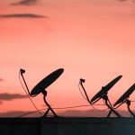 Antenas parabólicas em silhueta sobre um telhado, contra um céu alaranjado ao pôr do sol.