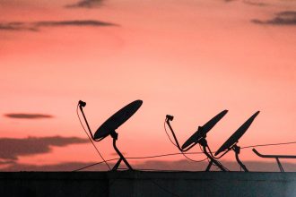Antenas parabólicas em silhueta sobre um telhado, contra um céu alaranjado ao pôr do sol.