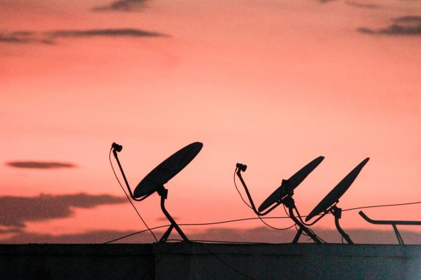 Antenas parabólicas em silhueta sobre um telhado, contra um céu alaranjado ao pôr do sol.