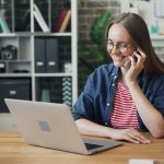 Mulher sorrindo enquanto usa o smartphone e o laptop em um escritório moderno.