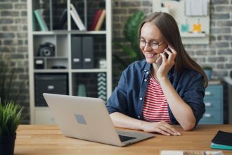 Mulher sorrindo enquanto usa o smartphone e o laptop em um escritório moderno.