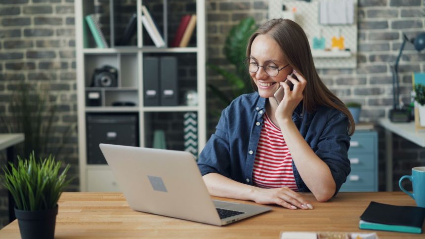 Mulher sorrindo enquanto usa o smartphone e o laptop em um escritório moderno.